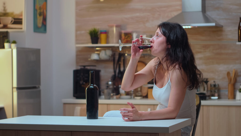 Woman drinking red wine alone in kitchen, illustrating alcoholism and health consequences